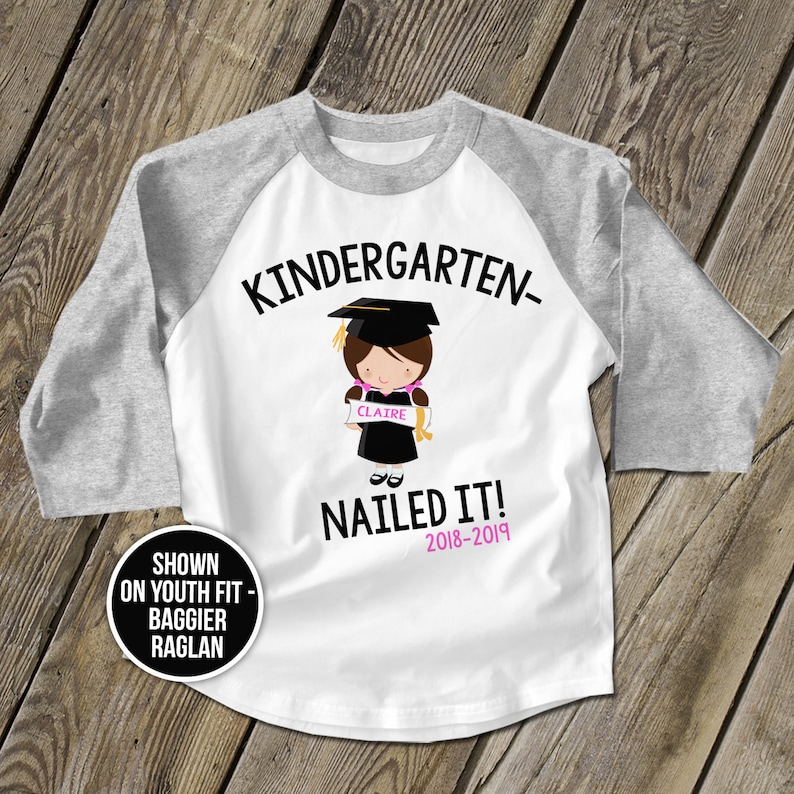 May include: White and grey youth baseball t-shirt with the text "Kindergarten- Nailed It! 2018-2019" and a cartoon illustration of a girl wearing a graduation cap and gown holding a scroll that says "Claire".