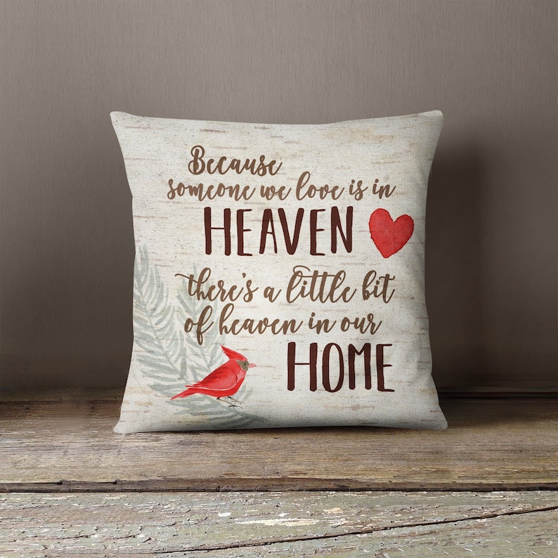 May include: A decorative throw pillow with a white background and brown text that reads "Because someone we love is in HEAVEN there's a little bit of heaven in our HOME". A red heart and a red cardinal bird are also featured on the pillow.