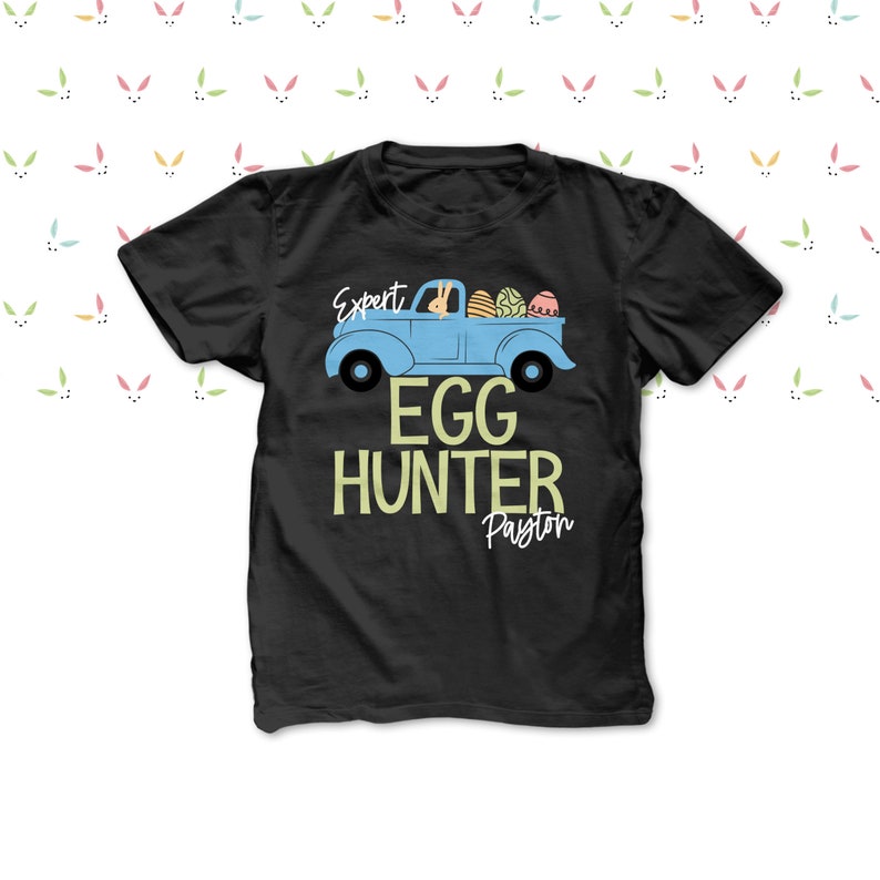 May include: Black t-shirt featuring a light blue truck graphic with Easter eggs. The text on the shirt reads "Expert Egg Hunter Payton". The design includes a bunny in the truck and a bunny ears pattern.