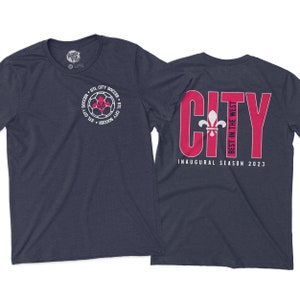 May include: A navy blue t-shirt with a white soccer ball and fleur-de-lis graphic on the front. The back of the shirt has the word "CITY" in pink with the text "BEST IN THE WEST" and "INAUGURAL SEASON 2023" in white.