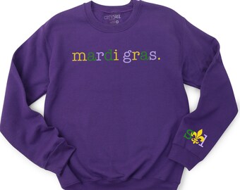May include: Purple sweatshirt with the text "mardi gras." in green, yellow, and purple lettering. The left sleeve has a small embroidered fleur-de-lis in green, yellow, and purple.