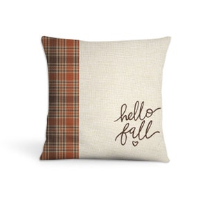 May include: A beige throw pillow with a brown and orange plaid border and the text "hello fall" in brown script with a small heart.