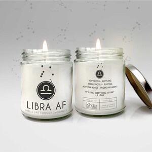 May include: A glass candle jar with a silver lid. The candle is white and has a black Libra zodiac symbol and the text "LIBRA AF" and "SMELLS LIKE CONFLICT AVOIDANCE". The jar also has the text "TOP NOTES - WAFFLING, MIDDLE NOTES - FLIRTING, BOTTOM NOTES - PEOPLE-PLEASING, "IT'S FINE, EVERYTHING IS FINE" - A LIBRA, 100% soy wax, weight: 9 oz, hand poured in Kirkwood, Missouri".