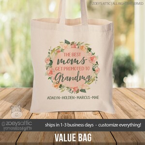May include: A white canvas tote bag with a floral wreath design. The wreath has pink and peach flowers and green leaves. The text in the center reads "The Best Moms Get Promoted to Grandma". Below the wreath, the text reads "Adalyn Holden Marcus Mae".