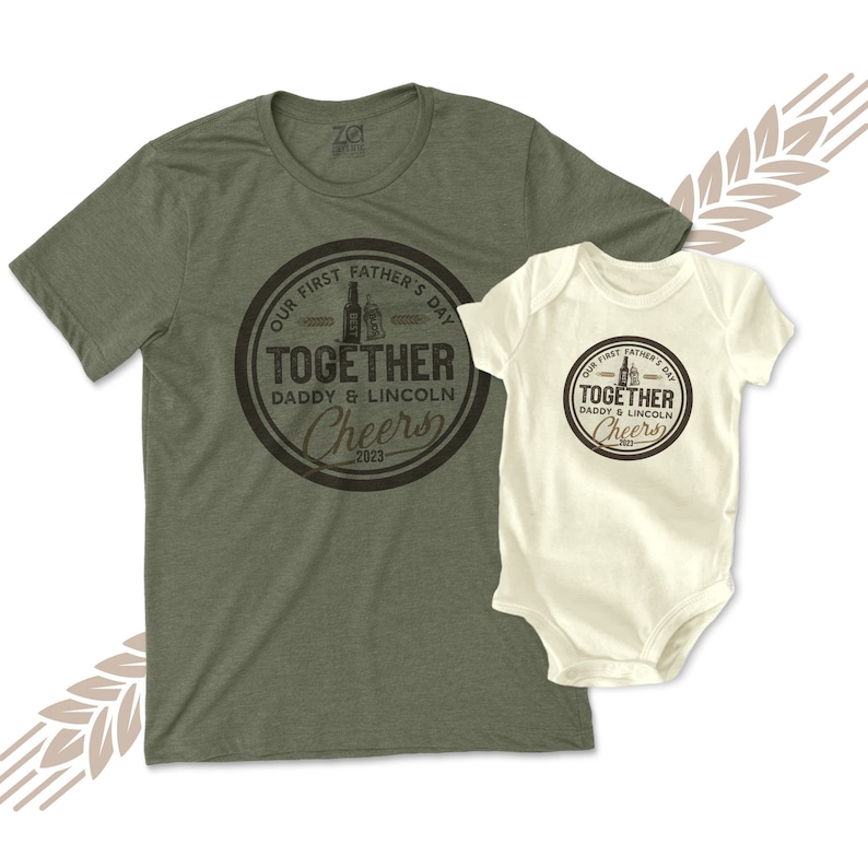 May include: A green t-shirt with the text "Our First Father's Day Together Daddy & Lincoln Cheers 2023" in a brown circle. A white baby onesie with the same text in a brown circle.