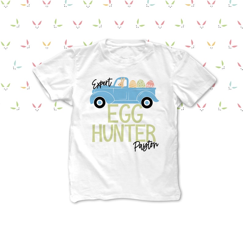 May include: White t-shirt with a light blue truck design carrying Easter eggs. The text reads "Expert Egg Hunter Payton". The background features a pattern of colourful bunny ears.