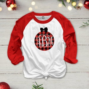 Op de afbeelding: Witte lange mouwen baseball shirt met rode mouwen. De voorkant van het shirt heeft een zwart en rood buffalo plaid kerst ornament met een zwarte strik en het monogram "IBM".