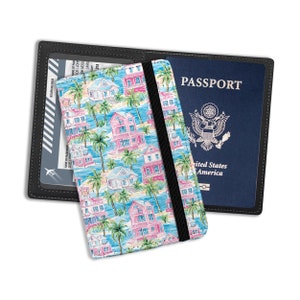 May include: A black leather passport holder with a blue and white patterned insert featuring pink houses and palm trees. The holder has a clear plastic window for a boarding pass and a black elastic band to keep it closed. The text "PASSPORT" is printed on the front of the holder.