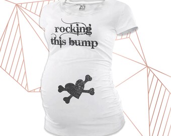 Può includere: T-shirt premaman bianco con la scritta "rocking this bump" in un font invecchiato. Sotto il testo c'è un cuore nero e un teschio con ossa incrociate. La maglietta ha un design aderente e lati arricciati.