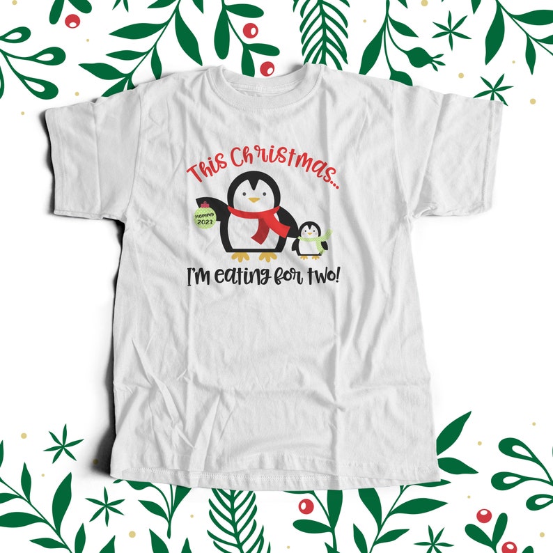 May include: White t-shirt featuring a penguin graphic and the text "This Christmas... I'm eating for two!" The penguin is holding a small gift box with the text "Mommy 2023".