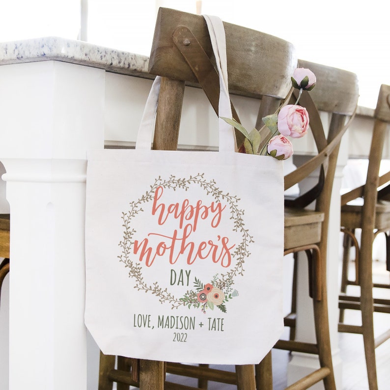 May include: White canvas tote bag with a floral wreath design and the text "happy mother's day" in pink. The text "love, madison + tate 2022" is printed below the wreath.