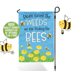 May include: A blue garden flag with the text "Please excuse the WEEDS we are feeding the BEES." The flag features illustrations of bees and yellow flowers. A black flag stand is also visible.