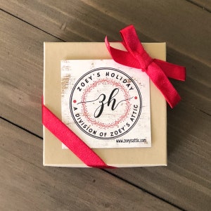 May include: A beige gift box with a red ribbon tied in a bow. The box has a white label with the text "ZOEY'S HOLIDAY A DIVISION OF ZOEY'S ATTIC zh www.zoeysattic.com"