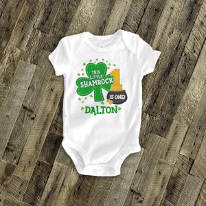 K&ouml;nnte beinhalten: Wei&szlig;er Baby-Strampler mit einem gr&uuml;nen Kleeblatt und einem goldenen Topf voller Gold. Der Text lautet "This little shamrock is one!" mit dem Namen "Dalton" darunter.
