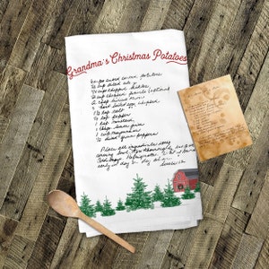 May include: A white tea towel with a handwritten recipe for "Grandma's Christmas Potatoes" on it. The recipe includes ingredients like potatoes, celery, pickles, and green peppers. The towel also features a red barn with a white door and a wreath, surrounded by green pine trees.