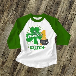 Puede incluir: Una camiseta de béisbol blanca y verde con un diseño de trébol y el texto "This Little Shamrock is Four!" y el nombre "Dalton".