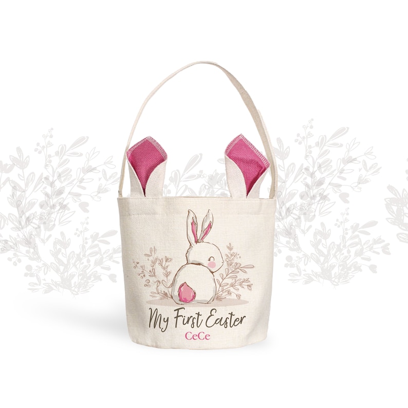 Puede incluir: Una cesta de Pascua de lona blanca con orejas de conejo rosas. La cesta tiene una linda ilustraci&oacute;n de conejo y el texto "My First Easter CeCe".