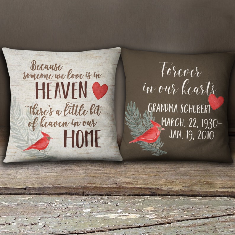 May include: Two decorative throw pillows with a brown background and a white background. The white pillow has a red heart and the text "Because someone we love is in HEAVEN there's a little bit of heaven in our HOME". The brown pillow has a red heart and the text "Forever in our hearts GRANDMA SCHUBERT MARCH. 22, 1930- JAN. 19, 2010".