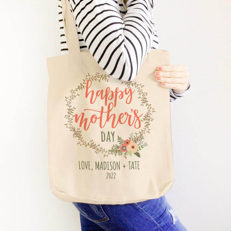 May include: A beige canvas tote bag with a floral wreath design and the text "happy mother's day" in red. The text "LOVE, MADISON + TATE 2022" is printed below the wreath.