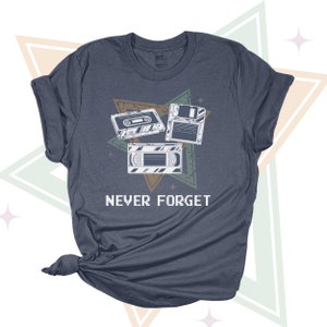 May include: A dark blue t-shirt with a vintage-style graphic of a cassette tape, floppy disk, and VHS tape. The graphic is surrounded by a starburst design and the text "NEVER FORGET".