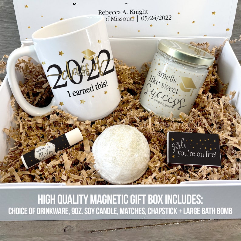 May include: A white gift box filled with graduation themed gifts. The box includes a white mug with gold stars and the text "2022 I earned this!" A white candle with gold stars and the text "smells like sweet success congrats, grad" A white bath bomb, a lip balm with the text "You're the Balm" and a matchbox with the text "girl, you're on fire!"