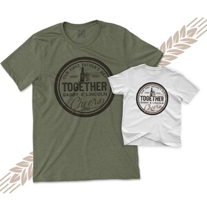 May include: A green t-shirt with a brown circle graphic that says "Our First Father's Day Together Daddy & Lincoln Cheers 2023". A white t-shirt with the same graphic but smaller is on top of the green t-shirt.