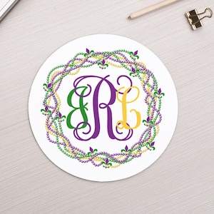 May include: A white round sticker with a purple, green, and gold beaded wreath with fleur-de-lis accents surrounding the monogram "BRL".