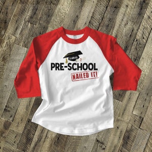 May include: White and red baseball style t-shirt with a graduation cap and diploma graphic. The text "PRE-SCHOOL NAILED IT!" is printed on the shirt. The name "Garrett" is on the graduation cap.