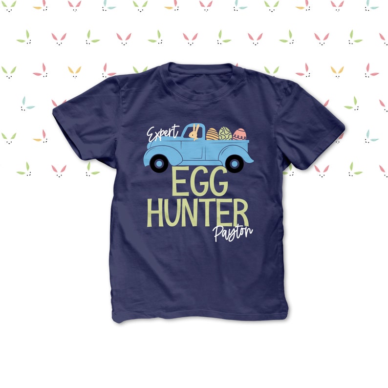 May include: Navy blue t-shirt featuring a light blue truck graphic filled with Easter eggs. The text "Expert Egg Hunter Payton" is printed below the truck. The background has a pattern of bunny ears.
