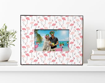 May include: A black picture frame with a white background and a pink flamingo pattern. The frame has a photo of a couple wearing sunglasses and holding a tropical drink.