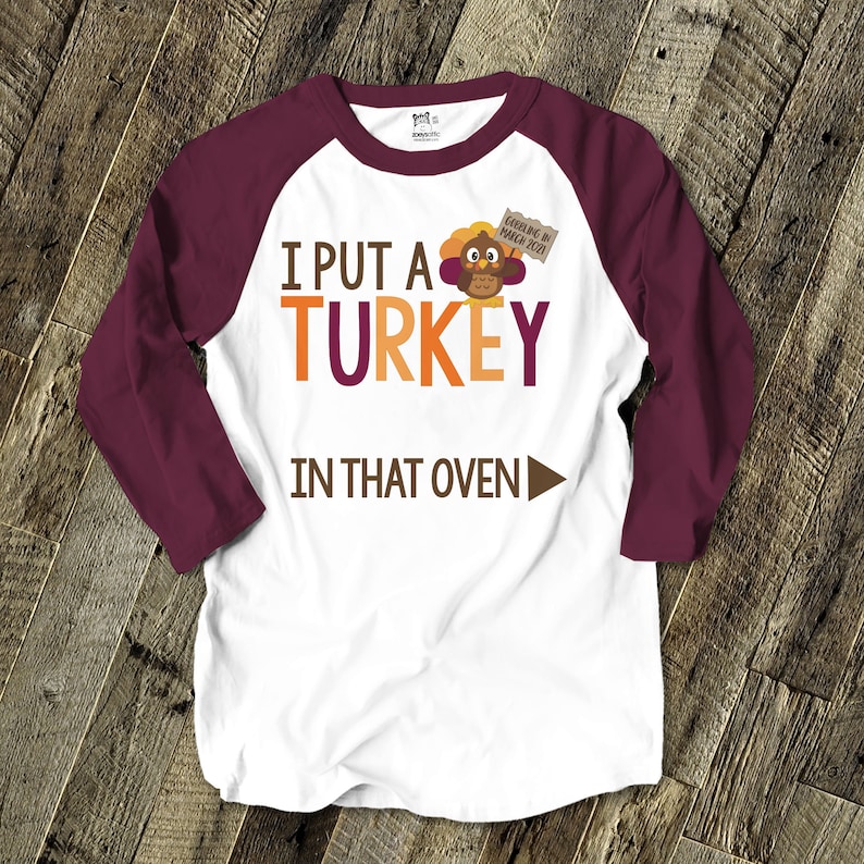 Pu&ograve; includere: Maglietta raglan bianca e bordeaux con la scritta "I PUT A TURKEY IN THAT OVEN" in arancione e marrone. Una grafica di un tacchino dei cartoni animati &egrave; sopra la parola "oven". La maglietta ha anche la scritta "Gobbling in March 2021".