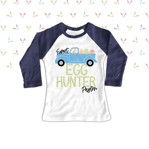 May include: White and navy blue raglan shirt with a light blue truck graphic carrying Easter eggs and a bunny. Text reads "Expert Egg Hunter" and a name below. The shirt is designed for Easter.