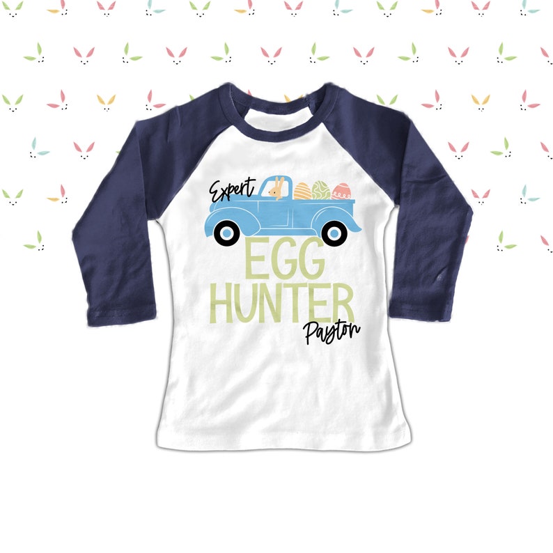 May include: White and navy blue raglan shirt with a light blue truck graphic carrying Easter eggs and a bunny. Text reads "Expert Egg Hunter" and a name below. The shirt is designed for Easter.
