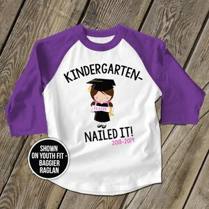 May include: White and purple baseball style t-shirt with the text "Kindergarten- Nailed It! 2018-2019" and a cartoon image of a girl wearing a graduation cap and gown holding a diploma that says "Claire".