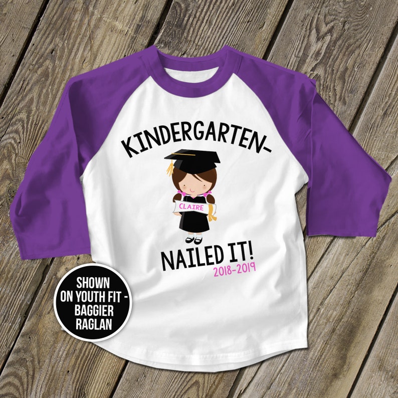 May include: White and purple baseball style t-shirt with the text "Kindergarten- Nailed It! 2018-2019" and a cartoon image of a girl wearing a graduation cap and gown holding a diploma that says "Claire".
