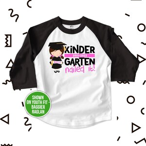 Op de afbeelding: Wit en zwart baseballshirt met een roze afbeelding van een meisje dat een afstudeerpet draagt en een diploma vasthoudt. De tekst "Kinder 2021-2022 Garten nailed it!" staat op het shirt gedrukt.