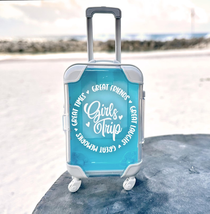 May include: A small blue suitcase with white wheels and a handle. The suitcase has a white circle sticker with the text "Girls Trip" and the words "Great Friends", "Great Times", "Great Memories", and "Great Laughs" around the circle.