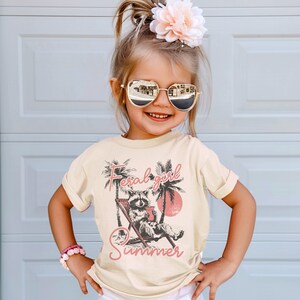 May include: A young girl wearing a light beige t-shirt with a graphic of a raccoon sitting in a lounge chair with the text "Feral girl summer" in pink lettering.