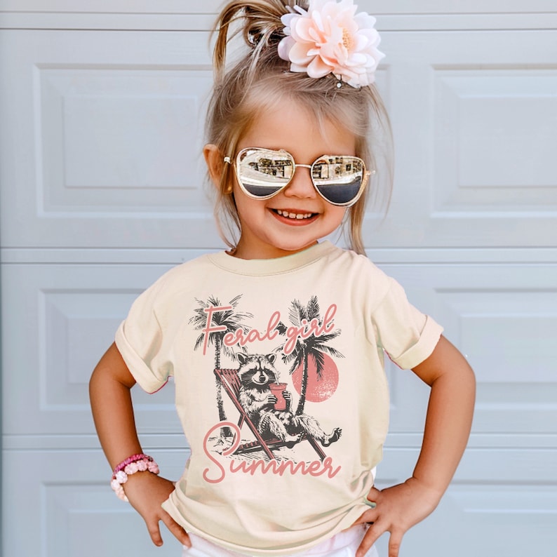 May include: A young girl wearing a light beige t-shirt with a graphic of a raccoon sitting in a lounge chair with the text "Feral girl summer" in pink lettering.