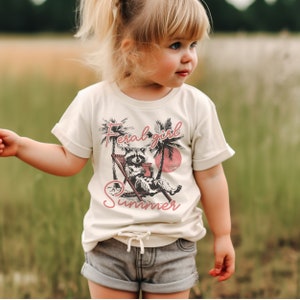 May include: A young person wearing a cream colored t-shirt with a graphic of a raccoon sitting in a lounge chair with the text "Feral girl summer" printed in pink.