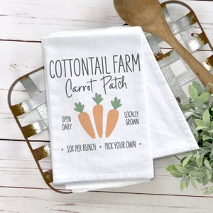 May include: White kitchen towel with the text "COTTONTAIL FARM Carrot Patch" in black. The towel also has the text "OPEN DAILY", "LOCALLY GROWN", and "10¢ PER BUNCH PICK YOUR OWN." There are three orange carrots with green tops.