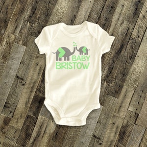 May include: White baby onesie with a green and grey elephant design. The text "BABY BRISTOW" is printed in green below the elephants.