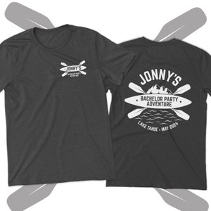 May include: A dark gray t-shirt with a white graphic design on the front and back. The front design says "Jonny's Bachelor Party Adventure" with two crossed paddles. The back design says "Jonny's Bachelor Party Adventure" with two crossed paddles, a mountain range, a lake, and the text "Lake Tahoe - May 2024".