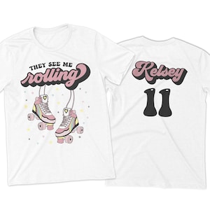 Puede incluir: Camiseta blanca con el texto "They See Me Rolling" en una fuente retro con un par de patines de ruedas rosas. La parte posterior de la camiseta tiene el nombre "Kelsey" en una fuente retro y el n&uacute;mero "11" en negro.