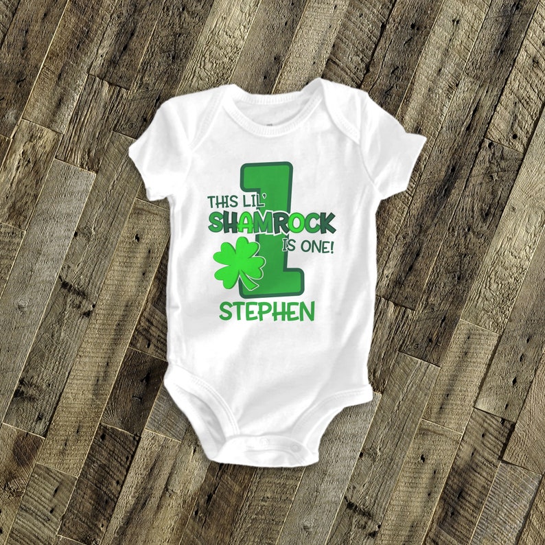 May include: White baby bodysuit with a green shamrock and the text "This lil' shamrock is one!" and a personalized name "Stephen".