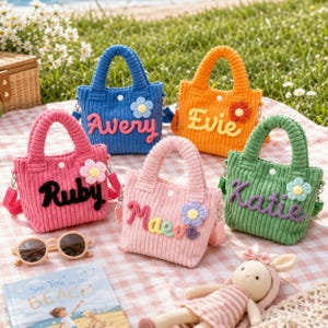 May include: Five small corduroy tote bags in blue, orange, pink, light pink, and green. Each bag features a personalised name in raised lettering and a small crocheted flower accent. The bags have two handles and a snap closure.