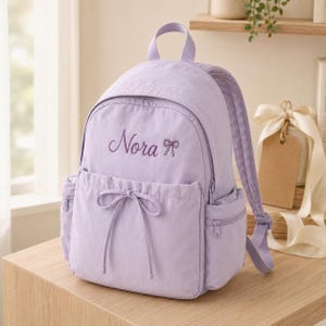May include: A light purple backpack featuring the embroidered name Nora and a small bow icon on the front pocket. The bag includes a top handle, adjustable shoulder straps, side zippered pockets, and a front drawstring closure with a tied bow.