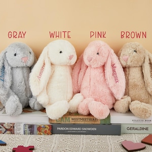 May include: Four plush bunny rabbits in gray, white, pink, and brown are displayed. Each bunny has long floppy ears and a pink nose. The ears are embroidered with names. The word "GRAY", "WHITE", "PINK", and "BROWN" are above each bunny.