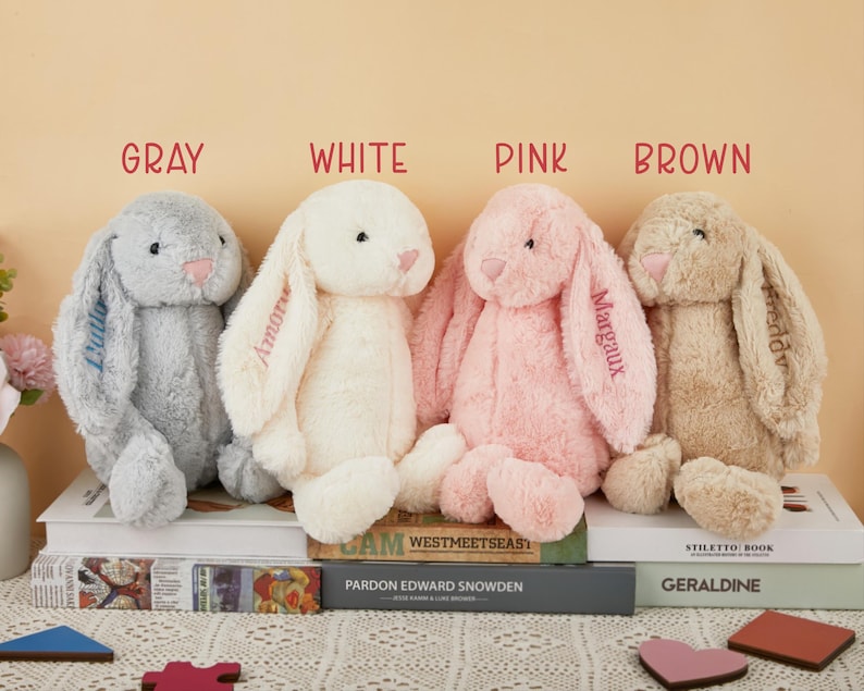 May include: Four plush bunny rabbits in gray, white, pink, and brown are displayed. Each bunny has long floppy ears and a pink nose. The ears are embroidered with names. The word "GRAY", "WHITE", "PINK", and "BROWN" are above each bunny.
