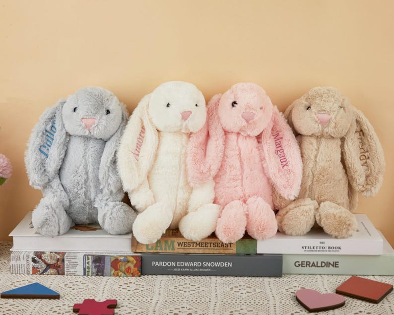 May include: Four plush bunny rabbits in gray, white, pink, and tan, each with embroidered names on their ears. The bunnies are sitting on a stack of books, with a neutral background.